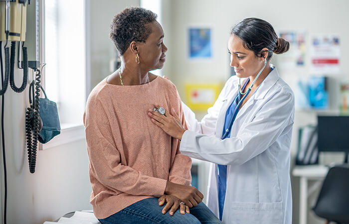 A senior woman of African decent sits up on an exam table during a routine check-up. She is sitting still and quiet as her female doctor listens to her heart.