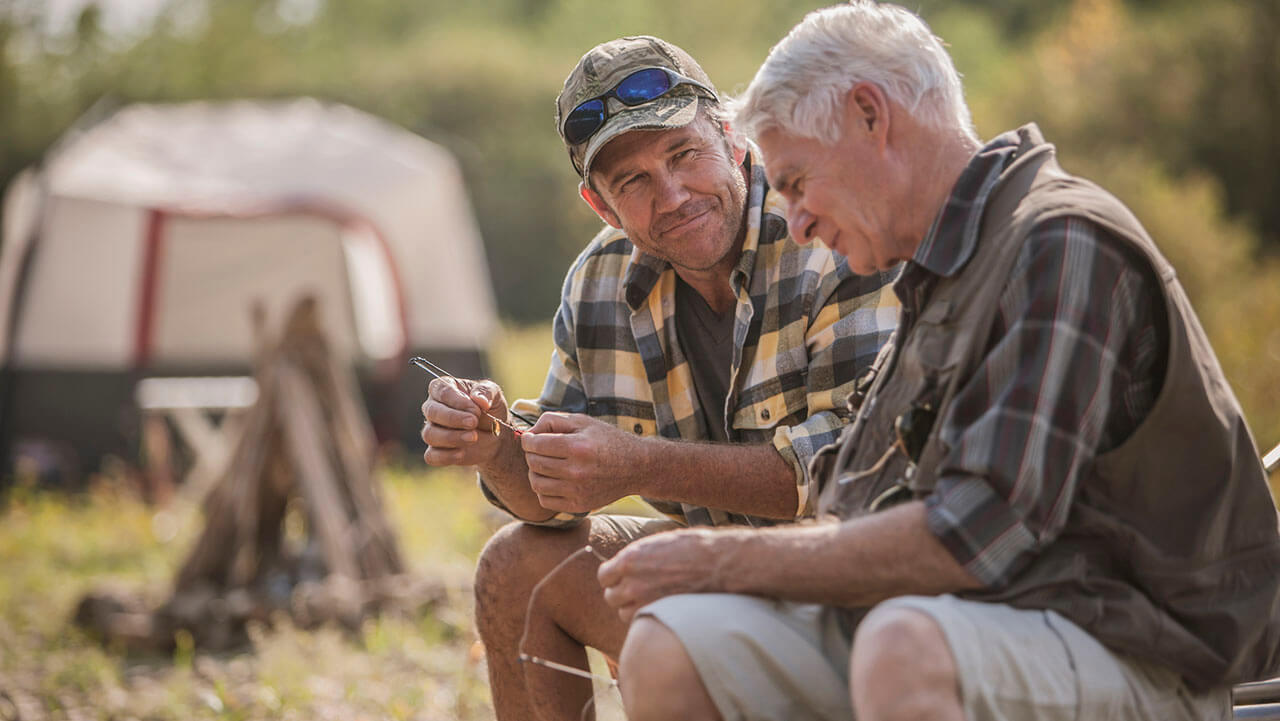2 men sitting outside in a camping setting; one man elderly, the other middle-age. 